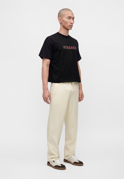 Άνδρας που φοράει μαύρο t-shirt Versace, παντελόνι εκρού και λευκά αθλητικά παπούτσια με καφέ και μαύρες λεπτομέρειες, στέκεται μπροστά σε απλό φόντο.