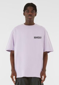Lys lilla oversized t-shirt lavet af bomuld, med et lille sort tekstgrafik på brystet. Korte ærmer og en afslappet pasform.