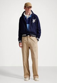 Casaco universitário acolchoado azul-marinho com riscas brancas e logótipo "P", camisa às riscas azuis em camadas, calças caqui de corte largo com remendos, ténis brancos.