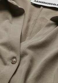 Camicia beige con bottoni, texture a coste, scollatura a V, chiusura con un singolo bottone e etichetta ARMEDANGELS visibile al colletto.
