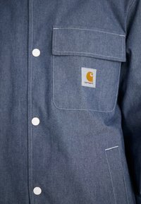Denim jacket in dark blue with white stitching. Features one chest pocket and a square logo patch with orange and white elements.