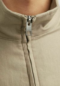 Bežna zip-up jakna s ovratnikom, gladke teksture, vidljivimi šivi in srebrnim kovinskim zadrgo. Ime blagovne znamke "Jack & Jones" na zadrgi.