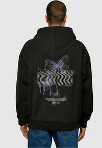 Sort hoodie med et stort grafisk motiv af gennemsigtige sommerfugle på bagsiden, med hvid stiliseret tekst nedenunder. Blødt materiale, afslappet pasform.