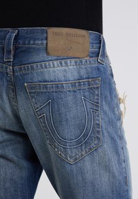 Blå denimjeans med en nötningseffekt, med bakfickor som har en signaturdesign med kurvad söm och en brun etikett med texten "True Religion."