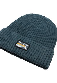 Bonnet en tricot à côtes couleur teal foncé avec un revers. Présente une patch noire avec des graphiques de montagnes et le texte "PROTEST" en blanc et orange.