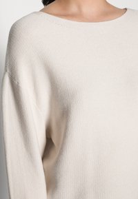 Lähikuva henkilöstä, joka on pukeutunut kermanväriseen, teksturoituun neulepuseroon, jossa on leveä, pyöreä pääntie.