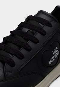 Μαύρο δερμάτινο αθλητικό παπούτσι με μαύρα υφασμάτινα κορδόνια και καρδιόσχημο μπαλώμα που γράφει «LOVE MOSCHINO» στο πλάι. Ορατή λευκή σόλα.