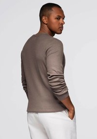Camisa de manga larga de color marrón oscuro y con canalé, con cuello redondo y puños ajustados, combinada con pantalones de color claro. La modelo tiene la espalda vuelta.