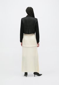 Blazer nero corto con finitura liscia, abbinato a una gonna plissettata color crema e stivaletti neri alla caviglia. La gonna presenta uno strato trasparente all'orlo.