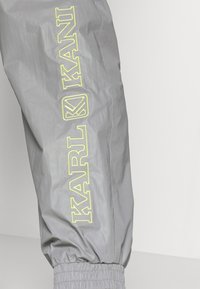 Graue Windbreaker-Hosen zeichnen sich durch elastische Bündchen und ein auffälliges gelbes "KARL KANI"-Logo an der Seite in einer markanten, modernen Schriftart aus.