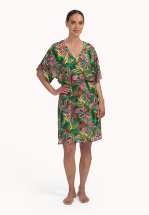 Vrouw staat op blote voeten en draagt een groene jurk tot op de knie met een tropische bloemenprint in roze en geel, korte mouwen en een V-hals.