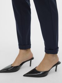 Črne lakaste usnjene mule z visokimi petami in koničastim dizajnom spredaj. Oblečene s temno modrimi hlačami z zavihanim robom, ki poudarjajo elegantne linije.