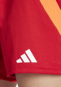 Rode atletische shorts met een gladde, gestructureerde oppervlakte. Heeft een oranje zijstreep en een wit Adidas-logo aan de zoom.