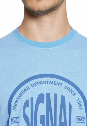 Mandens nakke og nedre del af ansigtet med let skægstubbe iført en lyseblå T-shirt med rund, mørkeblå tekst, der lyder "MENSWEAR DEPARTMENT SINCE 1967."