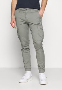 Homme portant un pantalon cargo slim gris clair avec des poignets élastiques, associé à des baskets blanches et une chemise bleu marine, se tenant devant un fond uni.