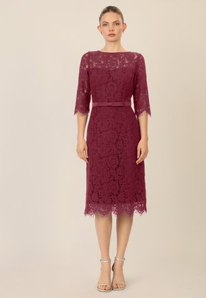 Burgunderfarbenes Spitzenkleid mit Dreiviertelärmeln, gewelltem Saum und einem passenden Gürtel. Ein Paar silberne Riemchenabsätze rundet den Look ab.
