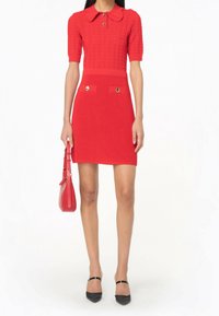 Robe rouge en tricot avec col, manches courtes et silhouette ajustée. Dotée de boutons dorés et de poches latérales. Chaussures à talons noires et sac à main.