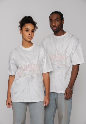 Zwei Erwachsene tragen weiße Batik-T-Shirts mit dem Aufdruck "Multiply" und stehen nebeneinander vor einem schlichten grauen Hintergrund.