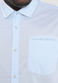 Jasnoniebieska koszula z krótkim rękawem wykonana z teksturowanej tkaniny, posiadająca klasyczny kołnierzyk, srebrne guziki oraz kieszeń na piersi z ozdobnym obszyciem.