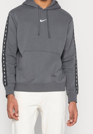 Mężczyzna w ciemnoszarej bluzie Nike z kapturem z logo na piersi i charakterystycznymi paskami na rękawach, ubrany w białe spodnie, stojący na tle białego tła.