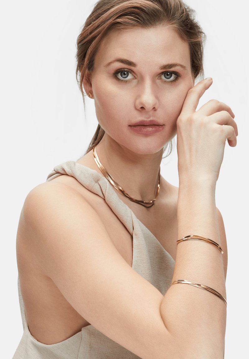 Gioielli in oro rosa comprendono una collana arrotolata e due braccialetti coordinati, indossati da un modello con un top strutturato e scollato.