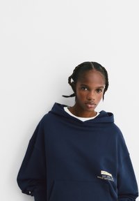 Námornícky modrá oversized mikina s kapucňou, vyrobená z jemnej bavlny, s klokankou a vyšitým logom na prednej strane. Jednoduché biele pozadie.