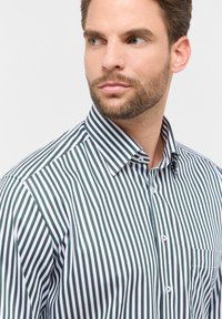 Man met kort bruin haar en baard, gekleed in een wit shirt met verticale donkergroene strepen, kijkt opzij tegen een effen achtergrond.