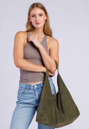 Joven mujer con cabello rubio largo, lleva una camiseta sin mangas marrón y vaqueros azul claro, sosteniendo un bolso grande de ante verde oliva para el hombro.