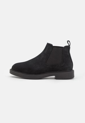 Sort ruskind Chelsea-støvle med elastisk sidepanel, trækflap ved hælen og lav gummisål på en ensfarvet hvid baggrund.