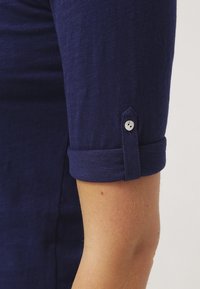 Mörkblå tröja med uppvikt ärm och knapppdetailj, tillverkad av en strukturerad och lätt tyg. Närbild av armen och skjortdesignen.