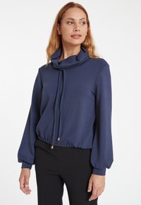 Navyfarbene Bluse mit Schalkragen, langen Ballonärmeln und einer Kordelzugdetail mit goldenen Akzenten, gefertigt aus glattem, weichem Stoff.