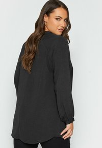 Överdimensionerad svart blus med långa ärmar, texturerad tyg, knäppte manschetter och rundad nederkant, med en lös passform och subtil drapering.