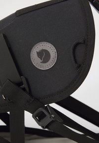 Lähetä lähellä oleva kuva mustasta pyöräilyrepusta, jossa on pyöreä Fjällräven-logo-patch ja säädettävät hihnat muovisilla soljilla.