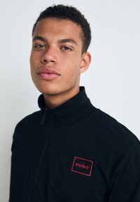 Svart zip-up jakke med høy krage, med rødt rektangulært logodetalj. Glatt stoff, tettsittende design.