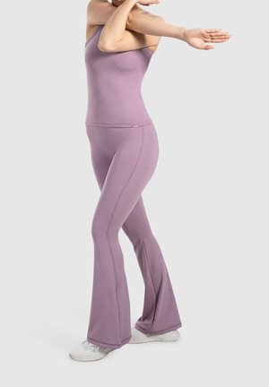 Žena natahující ruku přes hruď, oblečená v levandulovém tílku a rozšířených kalhotách, na nohou má bílé tenisky, na světlém pozadí.