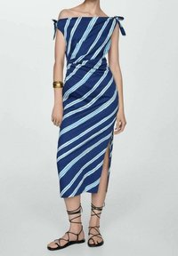 Robe décolletée en marine avec des rayures diagonales blanches et bleu clair. Comprend une fente latérale et des bretelles nouées. Associée à des sandales noires à lanières.