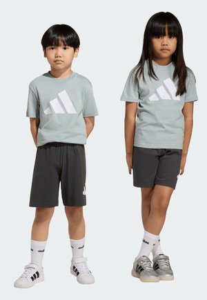 Due bambini indossano t-shirt grigie Adidas abbinate, pantaloncini neri, calzini bianchi Adidas e scarpe da ginnastica, in piedi davanti a uno sfondo semplice.