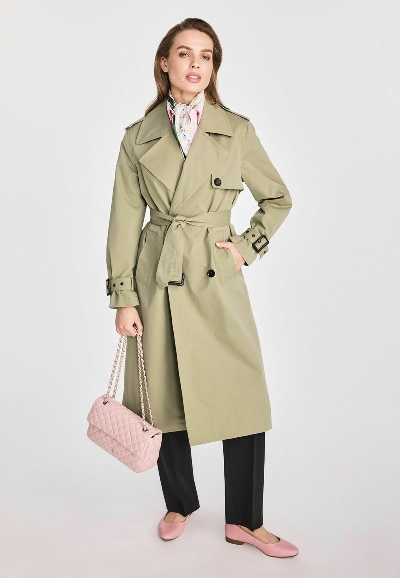 Jasnooliwkowy trencz z paskiem, czarnymi guzikami i podwójnymi kieszeniami; zestawiony z czarnymi spodniami oraz różową pikowaną torebką i balerinami.