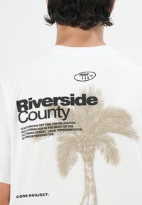 White cotton t-shirt featuring a palm tree graphic, "Riverside County" text, and additional descriptive text in black and brown.