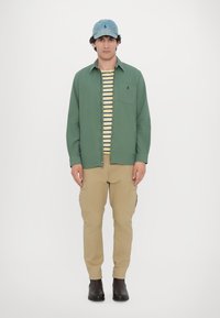 Camisa verde con cremallera sobre una camiseta de rayas amarillas y marinas, pantalones cargo beige, y una gorra de béisbol azul claro. Botas negras completan el look.