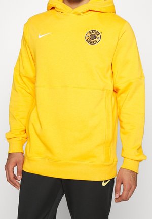 Gul Nike hættetrøje med Kaizer Chiefs-logo på brystet båret af en person i sorte Nike-bukser mod en hvid baggrund.