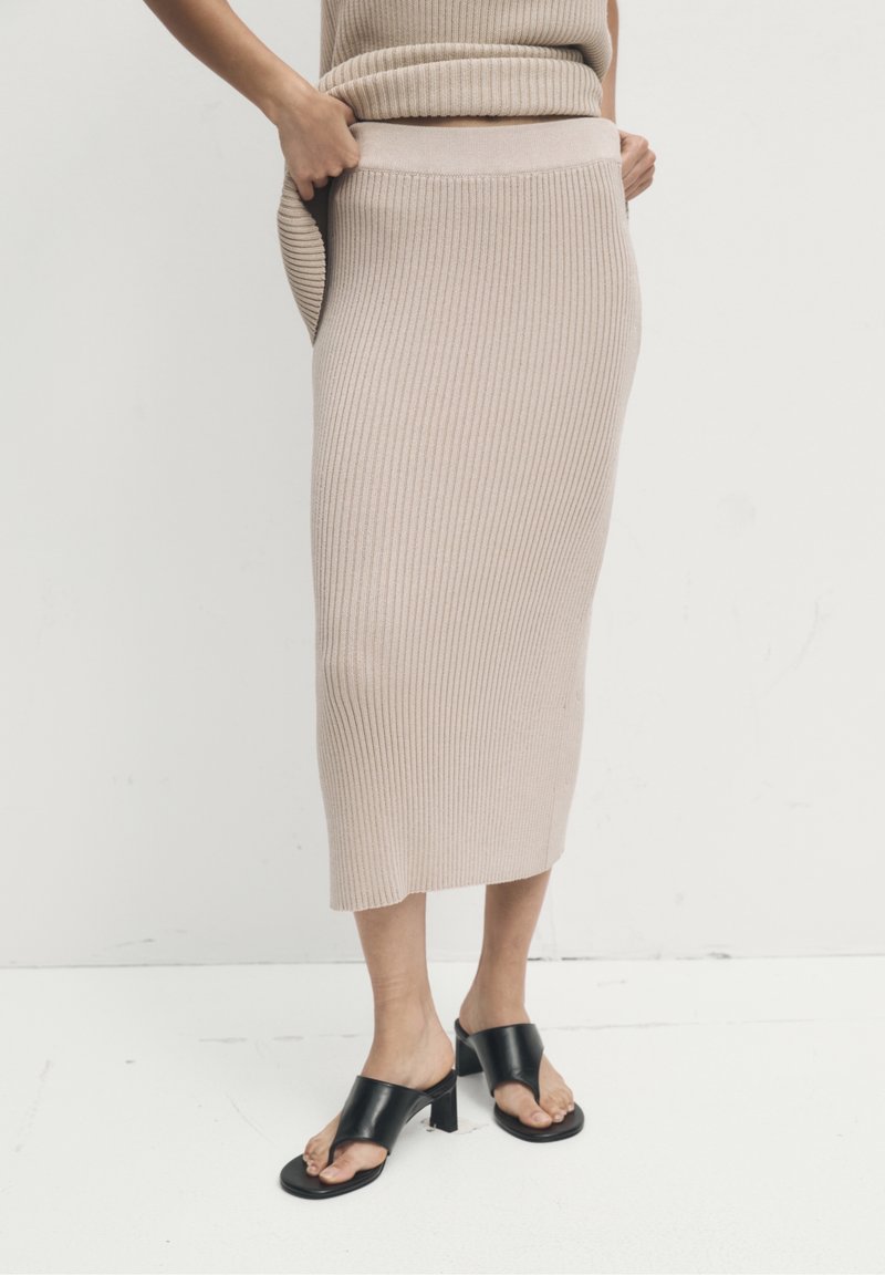 Personne portant une jupe midi en maille côtelée beige avec un haut assorti et des sandales à talons noires à bouts ouverts sur un sol blanc.