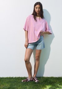 Prúžkované ružové a biele oversized tričko, svetlé denimbov�é šortky a hnedé topánky s kravským vzorom na slip-on. Pozadie trávy, neutrálny múr.