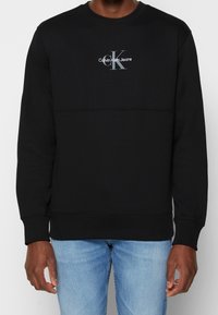 Svart sweatshirt med långa ärmar, rund halsringning, texturerat tyg, med en grå logotyp "Calvin Klein Jeans" på bröstet och en liten logotyp på handleden.