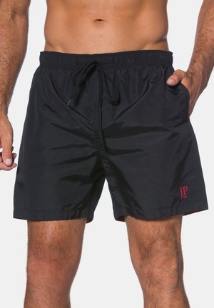 Mann trägt schwarze Badeshorts mit Kordelzug und rotem JP-Logo, steht mit den Händen in den Taschen vor einem hellen, einfarbigen Hintergrund.