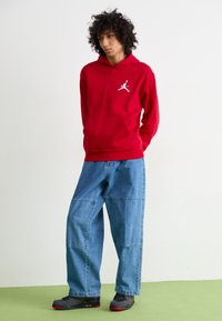 Rød bomulds-hoodie med et hvidt logo, parret med blå baggy denim-bukser. Fodtøj med sorte og røde accenter. Grønne gulve.