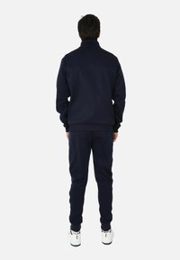 Tuta blu navy realizzato in tessuto morbido, con colletto alto, polsini a costine e pantaloni lunghi fino alla caviglia, abbinato a sneakers bianche.