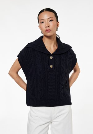 Kobieta ubrana w czarny bezrękawnik w splot warkocza z szerokim kołnierzem i dwoma guzikami, w połączeniu z białymi spodniami, stoi z rękami za plecami.