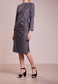 Robe grise texturée avec de longues manches, détail drapé sur le côté, longueur mi-mollet et fentes latérales, assortie à des sandales à talons blocs noires.