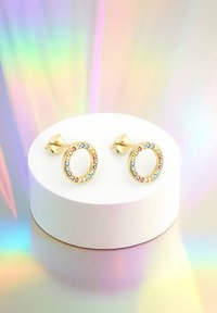 Orecchini circolari in oro con cristalli multicolori lungo il bordo, esposti su una piattaforma bianca con uno sfondo riflettente e iridescente.